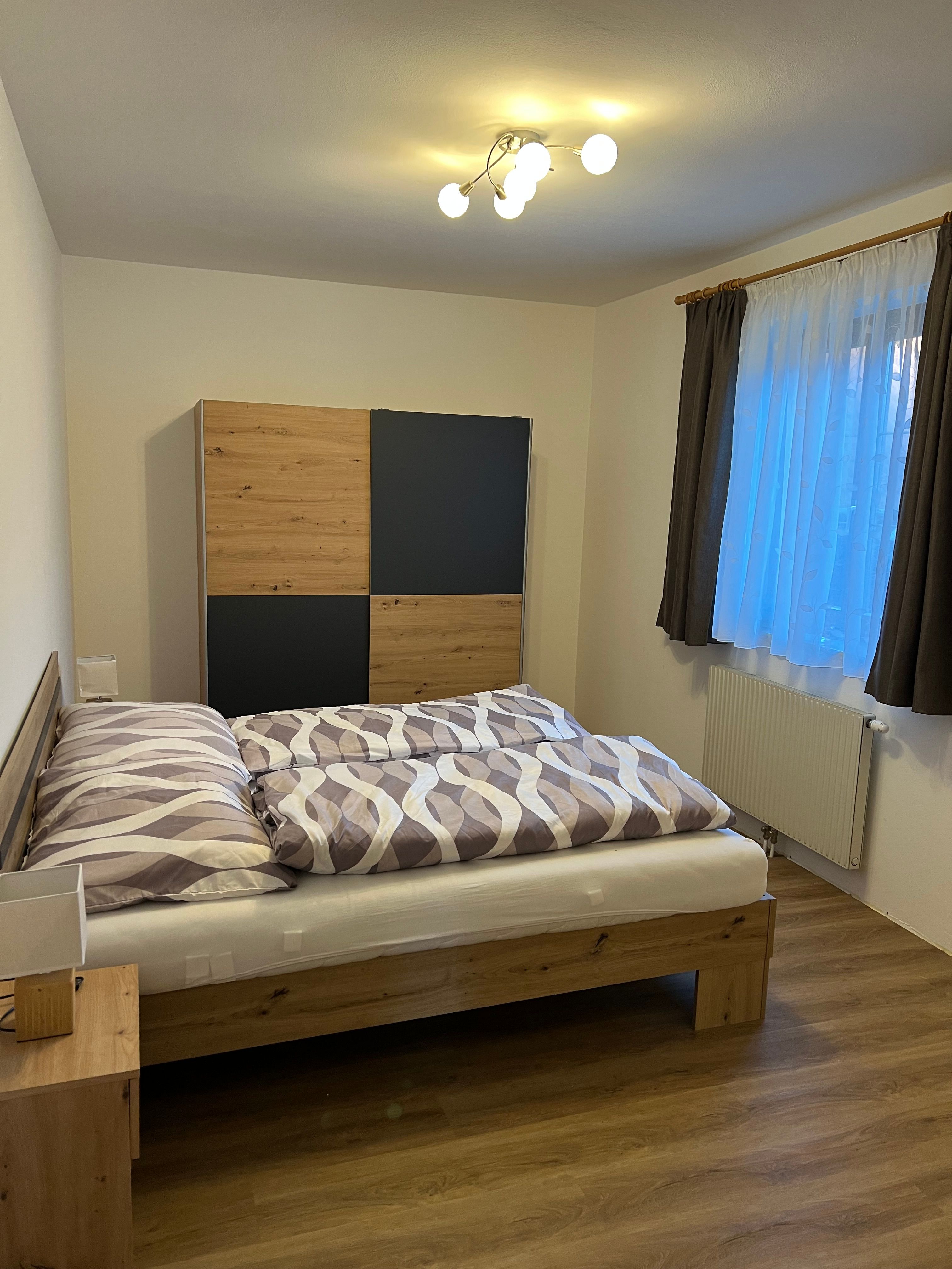 Útulný dvoulůžkový pokoj s dřevěnou postelí, šatní skříní a oknem se závěsy.