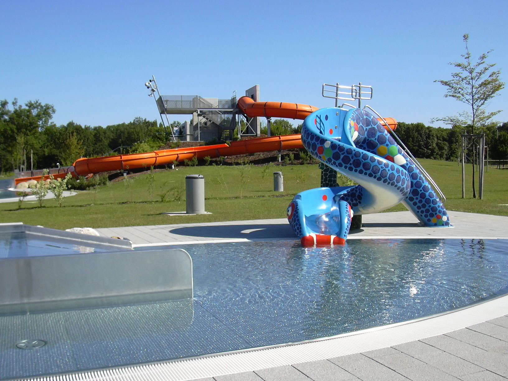 Pohled na aquapark s modrou dětskou skluzavkou a velkým oranžovým tobogánem v pozadí.