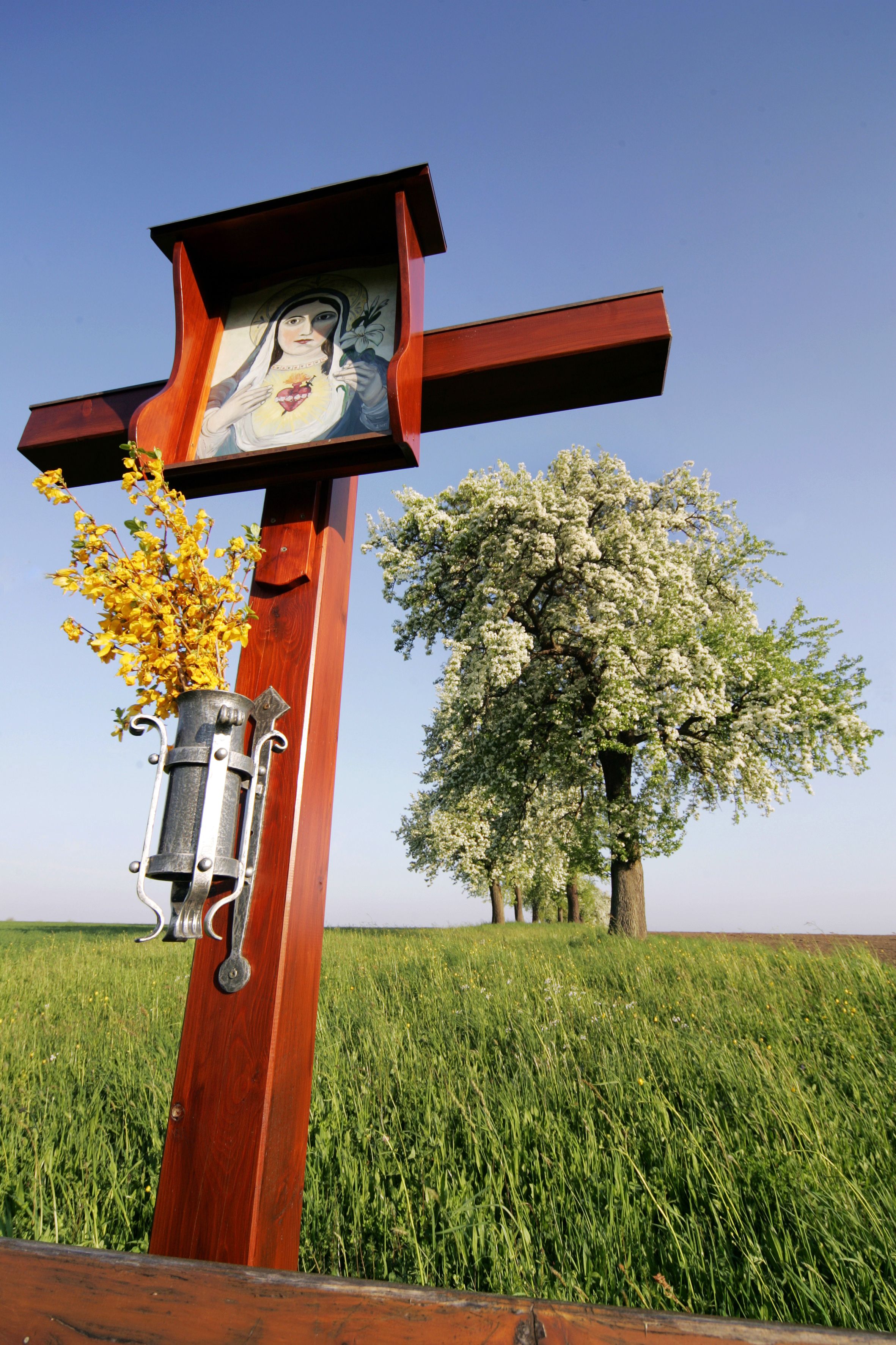 Dřevěný kříž u cesty s obrázkem Panny Marie a žlutými květy, v pozadí kvetoucí stromy na louce.