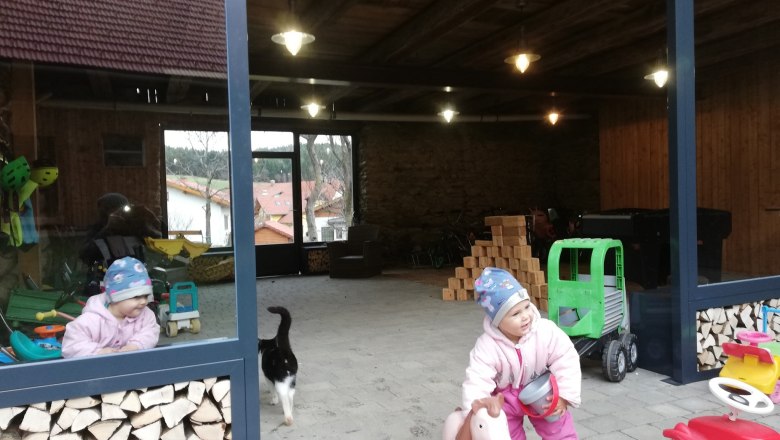 Dítě v zimním oblečení si hraje s koníkem v otevřené místnosti s hromadami dřeva a hraček. Kolem prochází kočka.