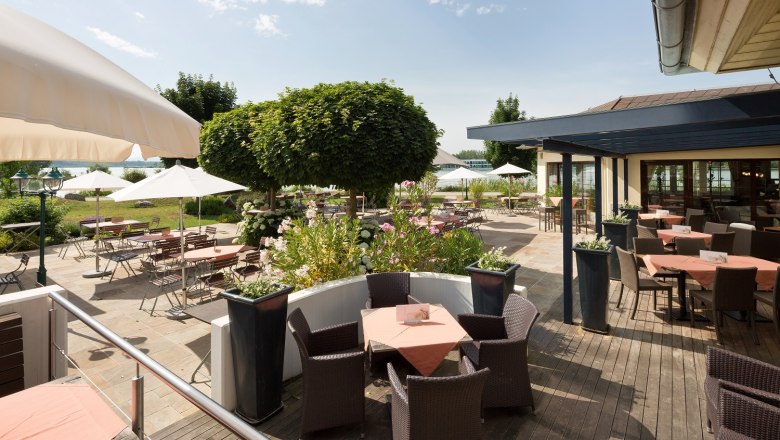 Sonnige Terrasse mit Tischen, St&uuml;hlen und Sonnenschirmen, umgeben von Pflanzen und B&auml;umen.