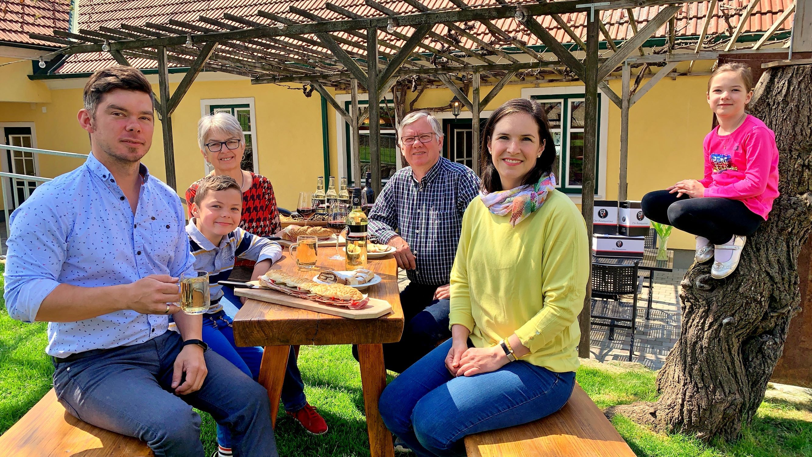 Rodina sedí venku u dřevěného stolu a vychutnává si jídlo. Dítě sedí na kmeni stromu. V pozadí je žlutý dům s pergolou.