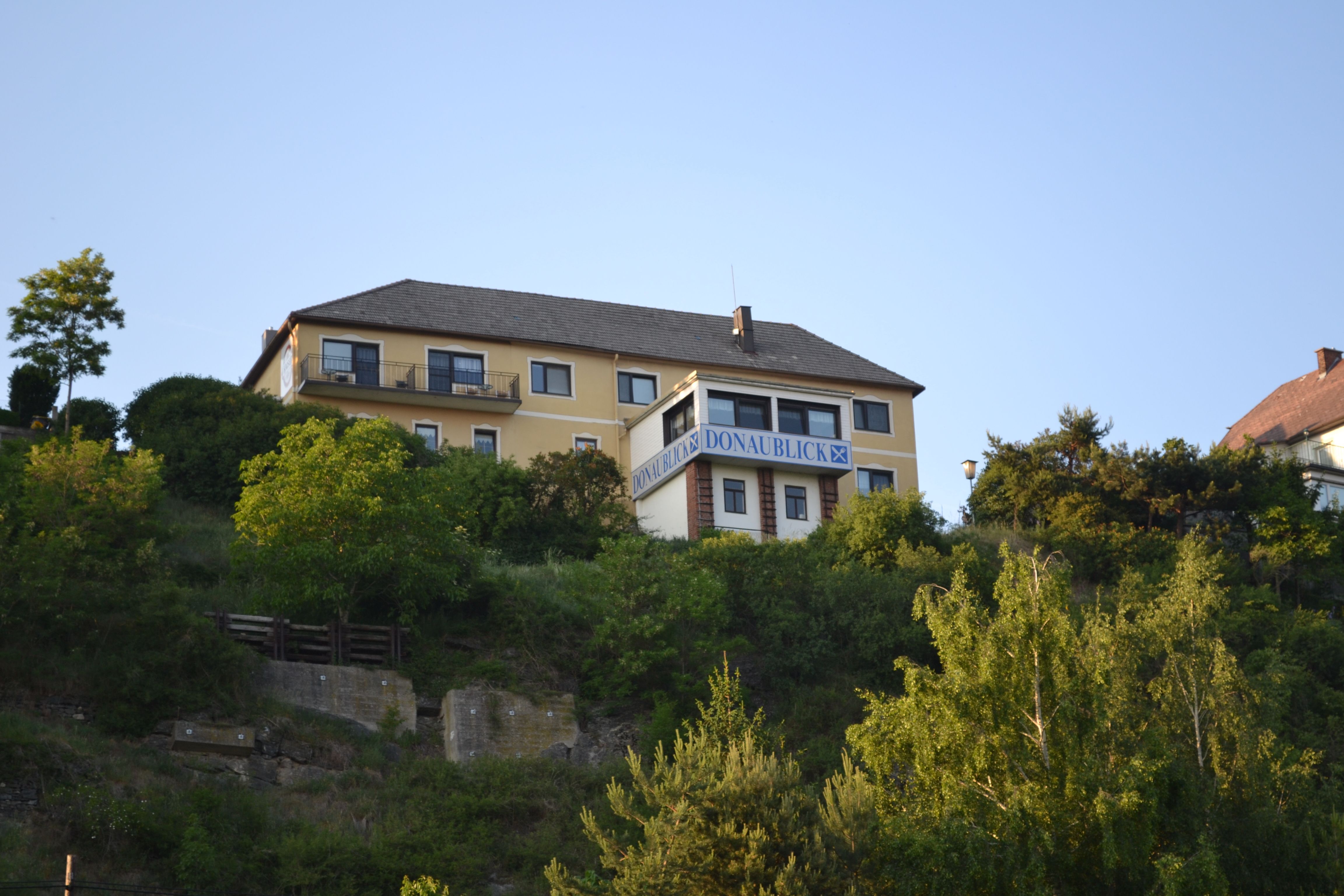 Žlutá budova s nápisem "Donaublick" na kopci, obklopená stromy a modrou oblohou.
