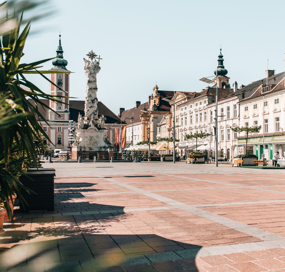 Die sonnendurchflutete Platzlandschaft lädt zum Verweilen ein, während die historischen Gebäude im Hintergrund eine charmante Kulisse bieten. Palmen und bunte Blumen verleihen dem Ort eine lebendige Atmosphäre, perfekt für entspannte Sommertage.