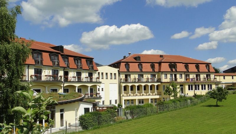 Kurhotel Moorbad Großpertholz s červenou střechou a balkony, obklopený zelenými loukami a stromy pod modrou oblohou.
