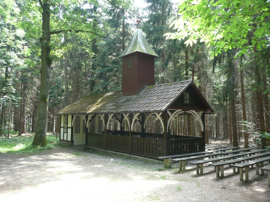 Malá kaple v lese s dřevěnými lavičkami venku.