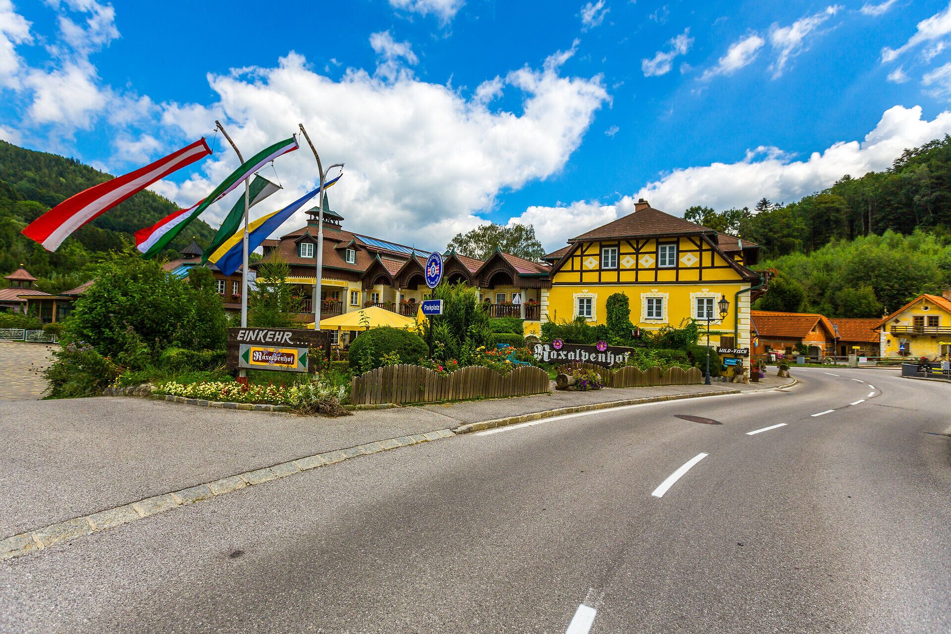 Raxalpenhof se nachází přímo u místní komunikace, před domem je modrá fontána, balkony a celou přední část domu lemují květiny a keře.
