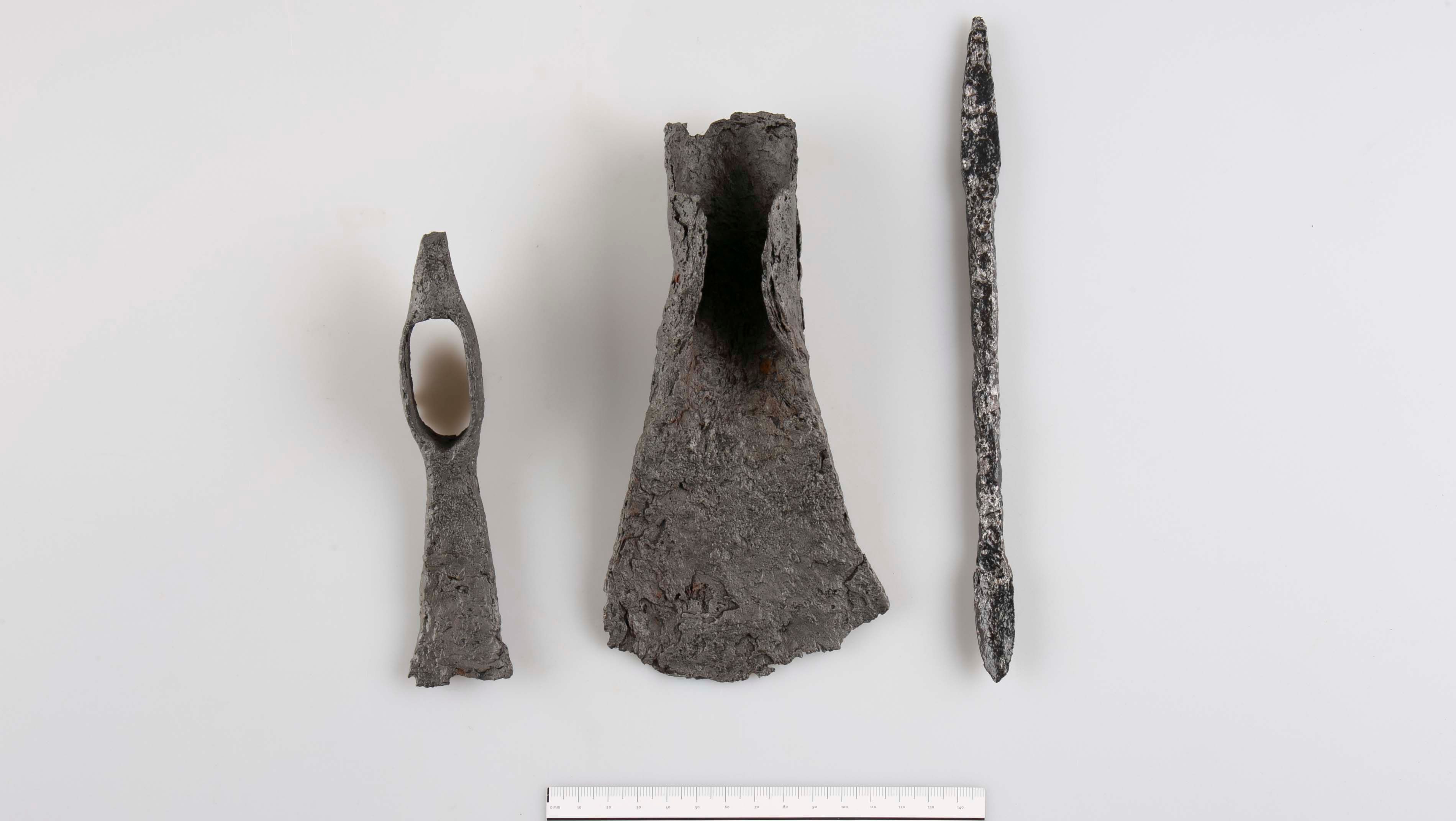 Tři archeologické kovové předměty na bílém pozadí se stupnicí ve spodní části.