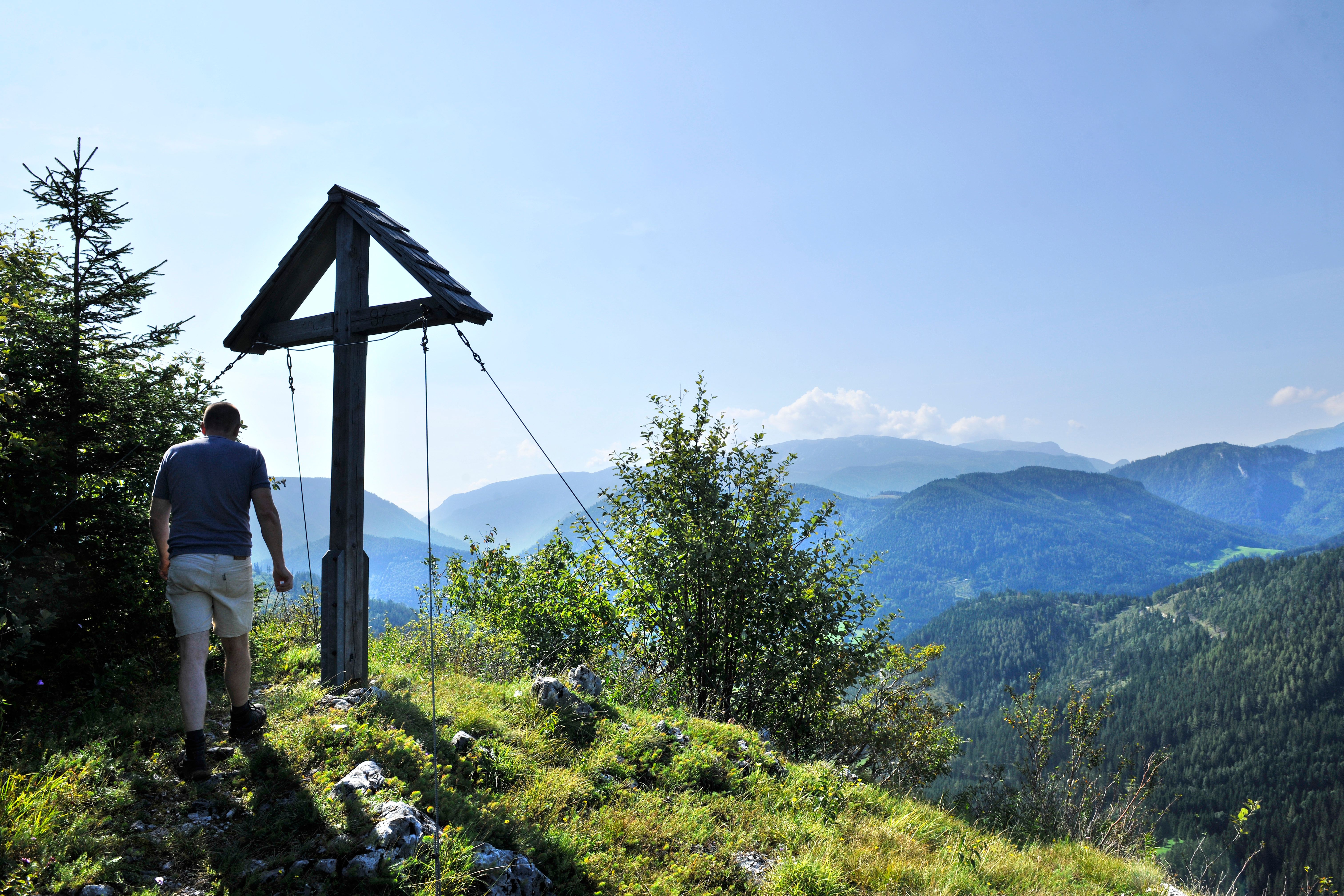 Turista stojí u vrcholového kříže s výhledem na zalesněné hory.