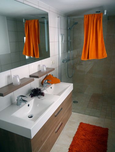 Moderní koupelna s dvojitým umyvadlem, velkým zrcadlem, sprchou a oranžovými akcenty.