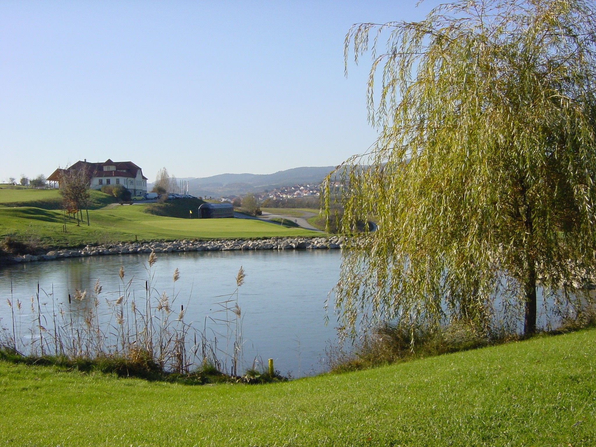 Golfové hřiště s rybníkem, stromy a budovou v pozadí.