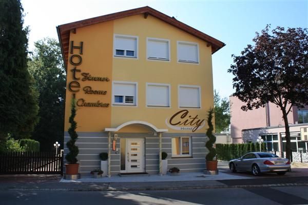 Žlutá budova s nápisem "City Hotel", obklopená stromy a zaparkovaným autem.