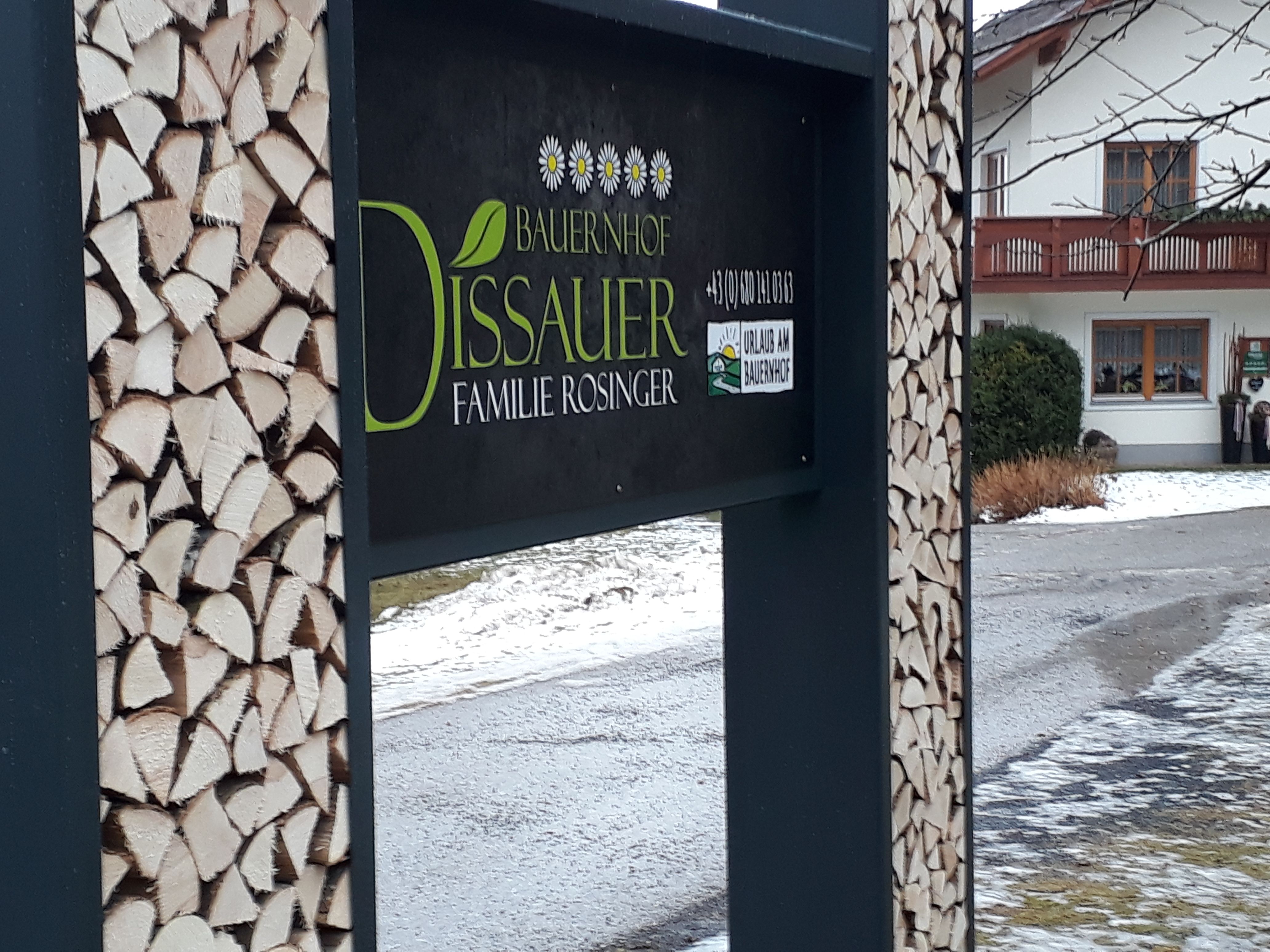 Znak farmy s hromadami dřeva a domem v pozadí.