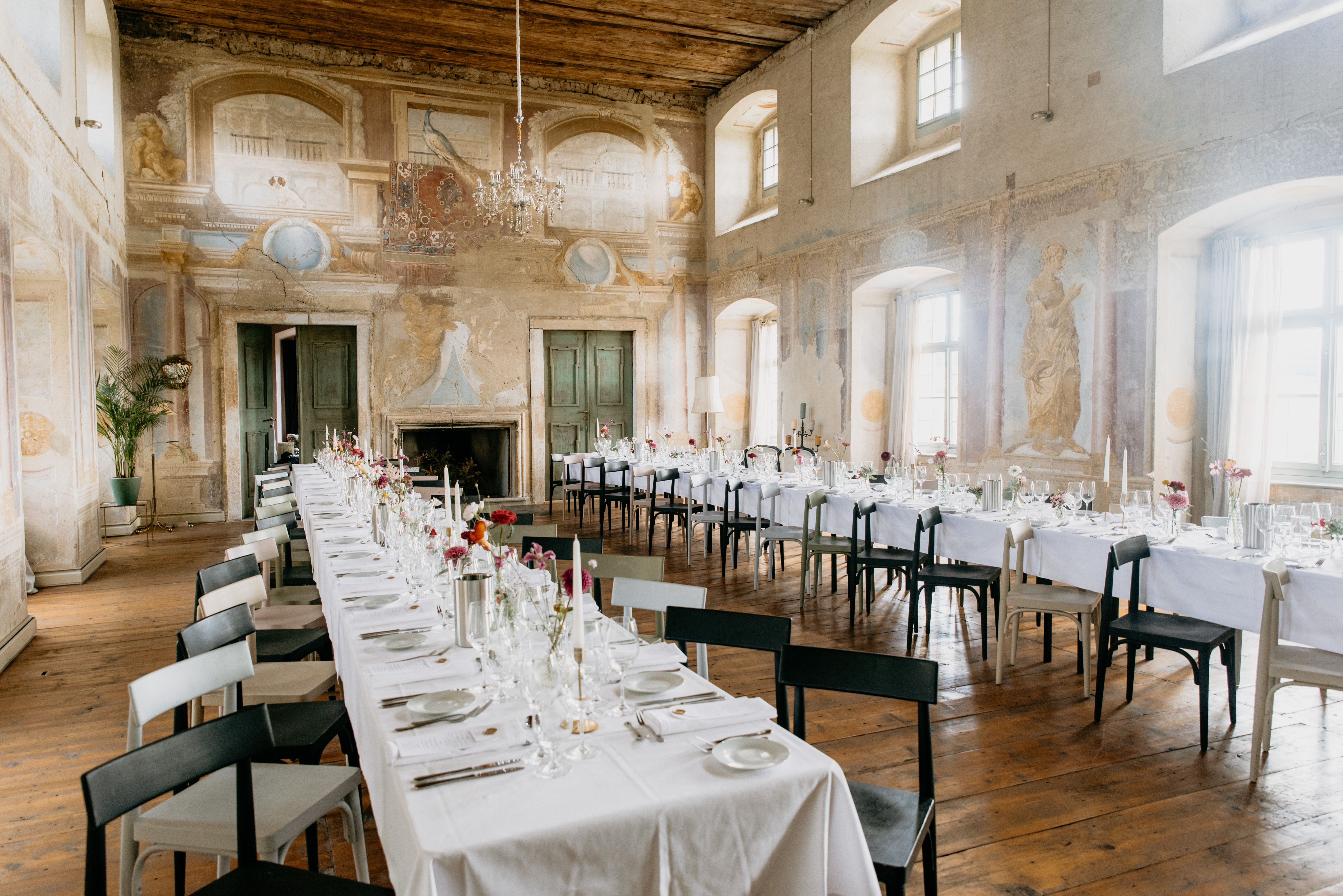 Elegantní hodovní sál s dlouhými prostřenými stoly, starožitnými nástěnnými malbami a lustry.