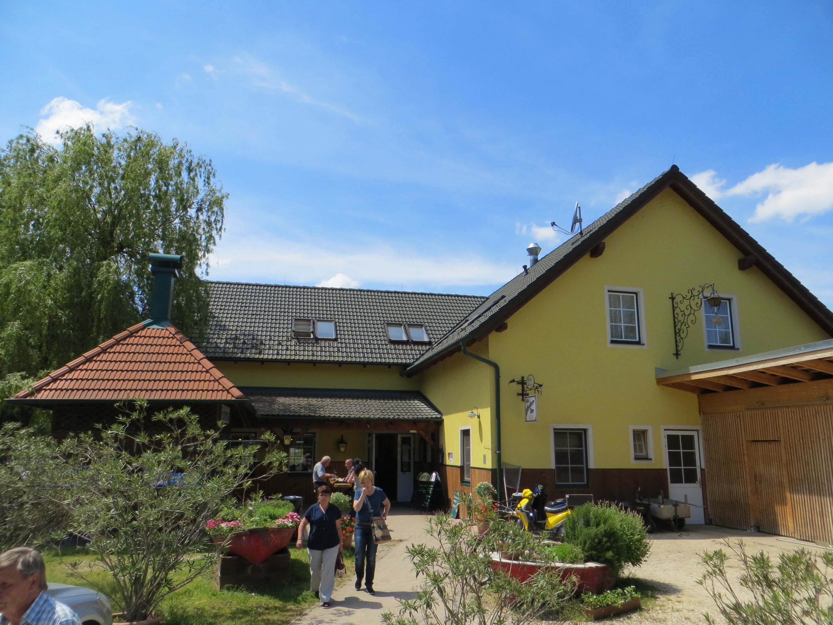 Žlutý hostinec s šedou střechou a zahradou v popředí.