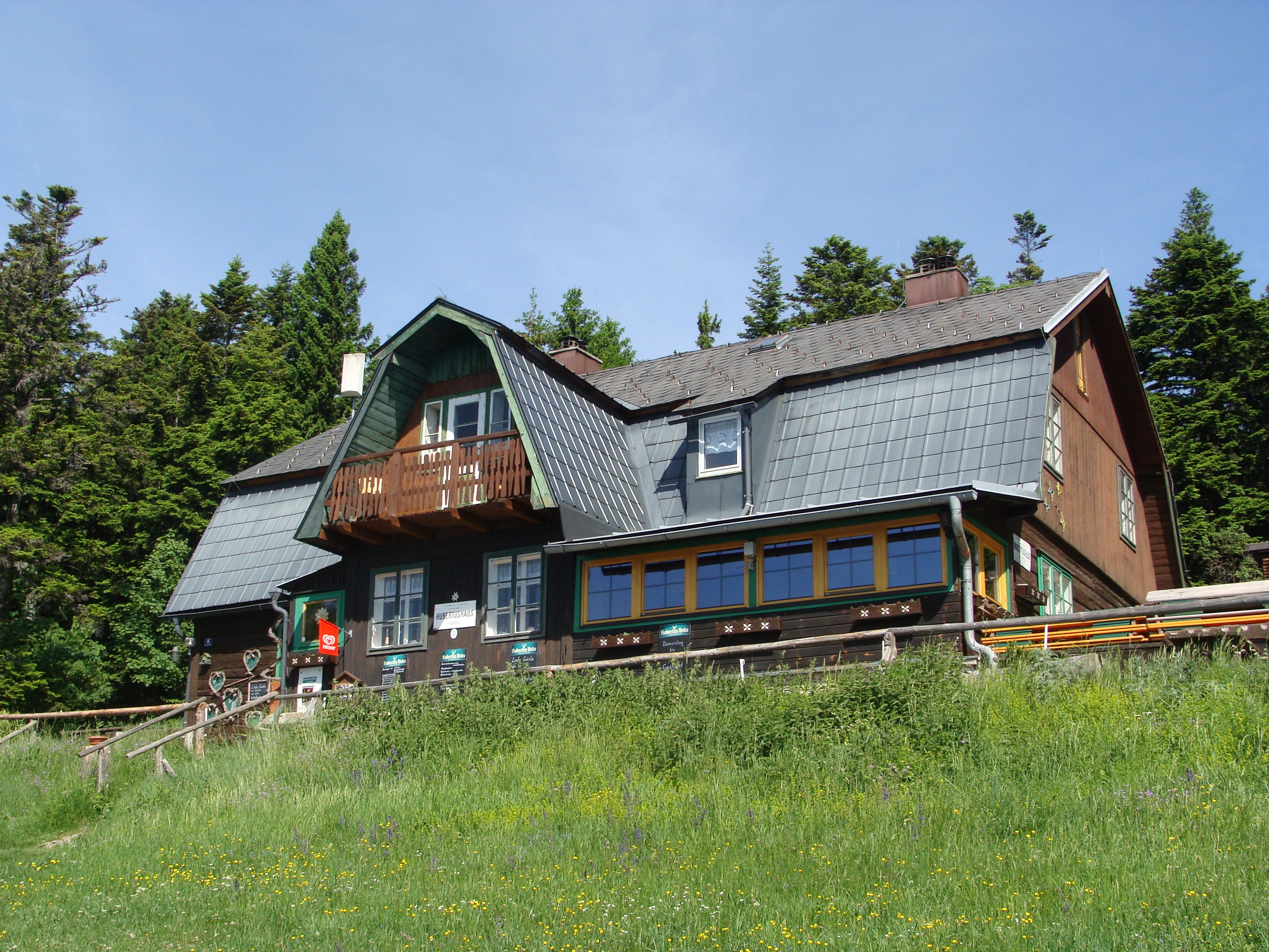 Tradiční horský dům s dřevěnou fasádou a šindelovou střechou, obklopený stromy a loukami.