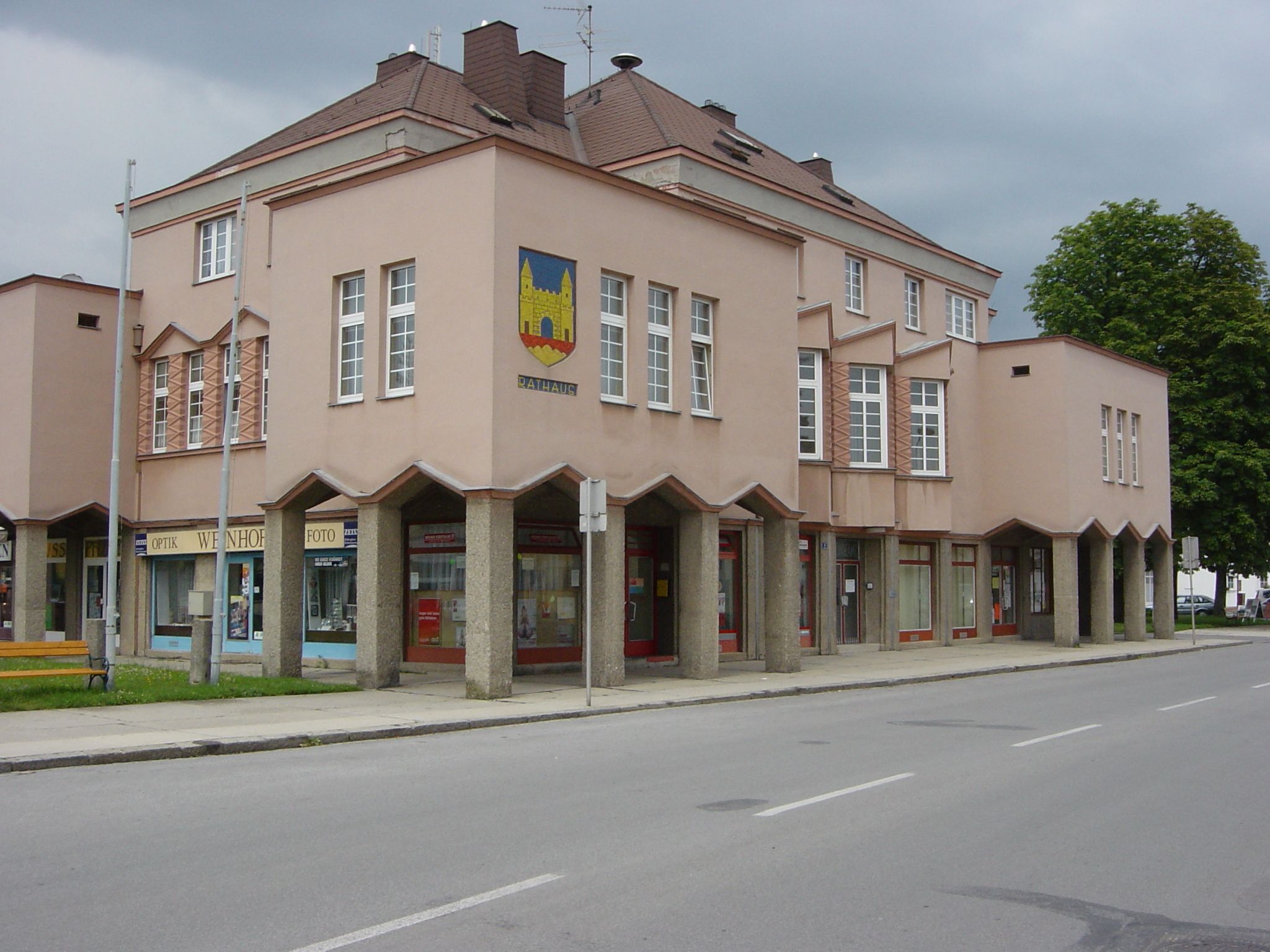 Moderní budova radnice s růžovou fasádou a velkými okny. Na nároží má erb a v přízemí obchody.