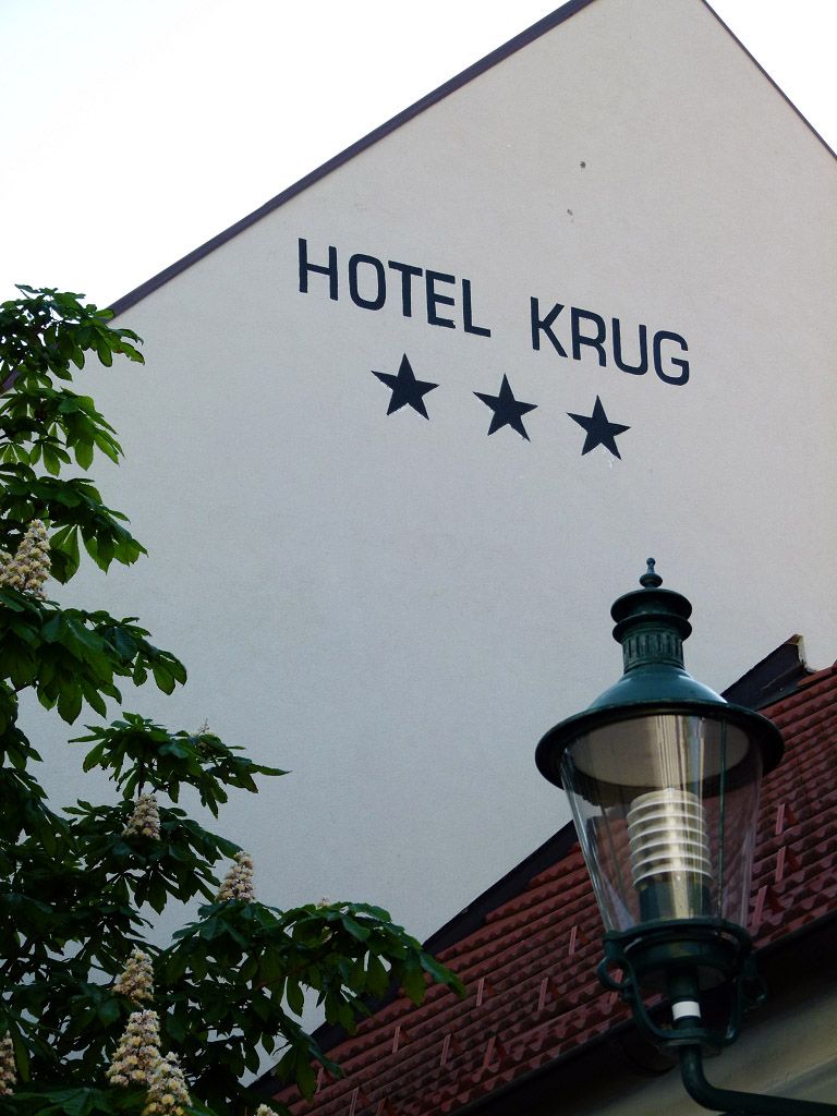 Fasáda hotelu Krug se třemi hvězdami, stromem a pouliční lampou.