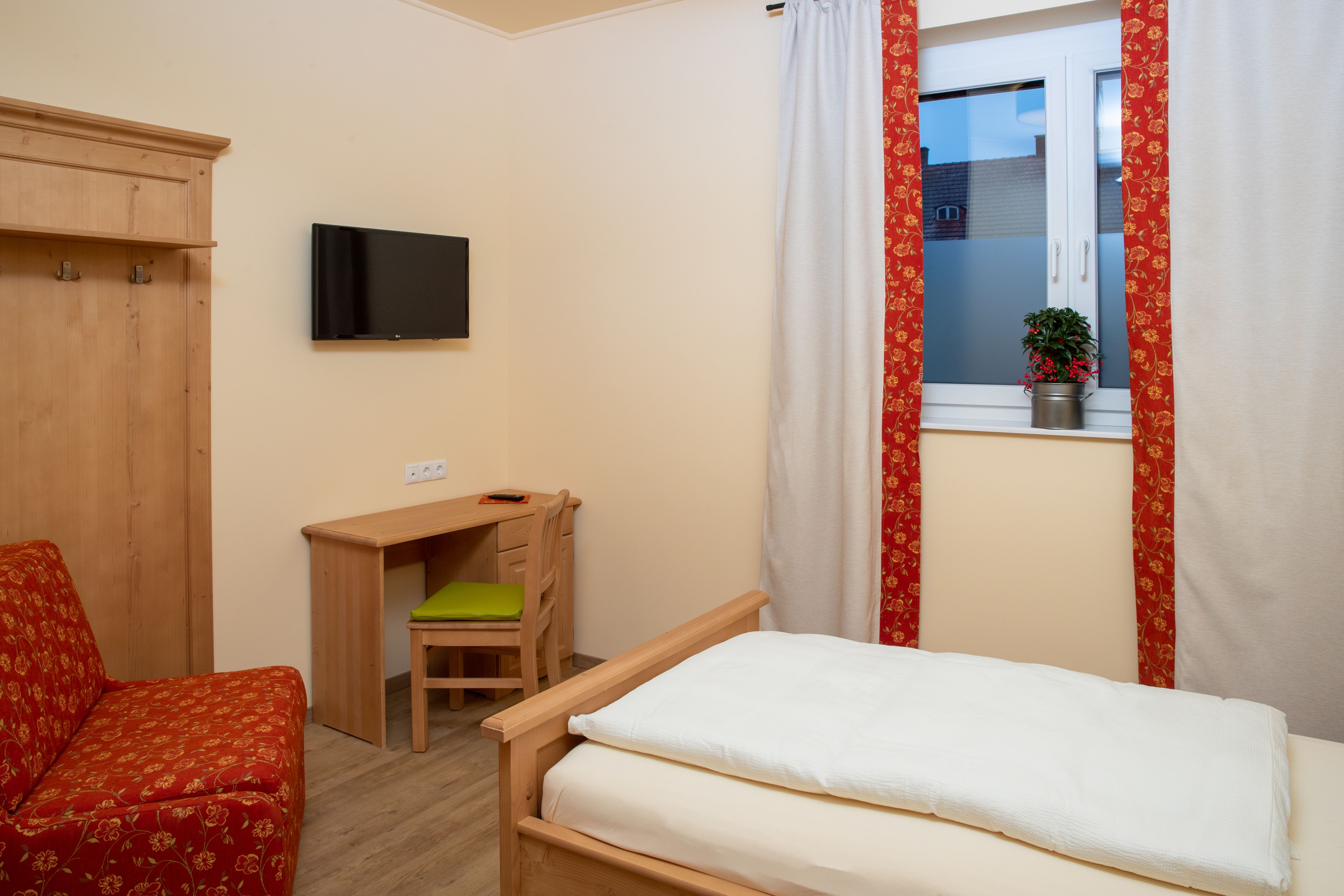 Útulný jednolůžkový pokoj s dřevěným nábytkem, postelí, psacím stolem a televizí na stěně. Okno se závěsy a rostlinou.