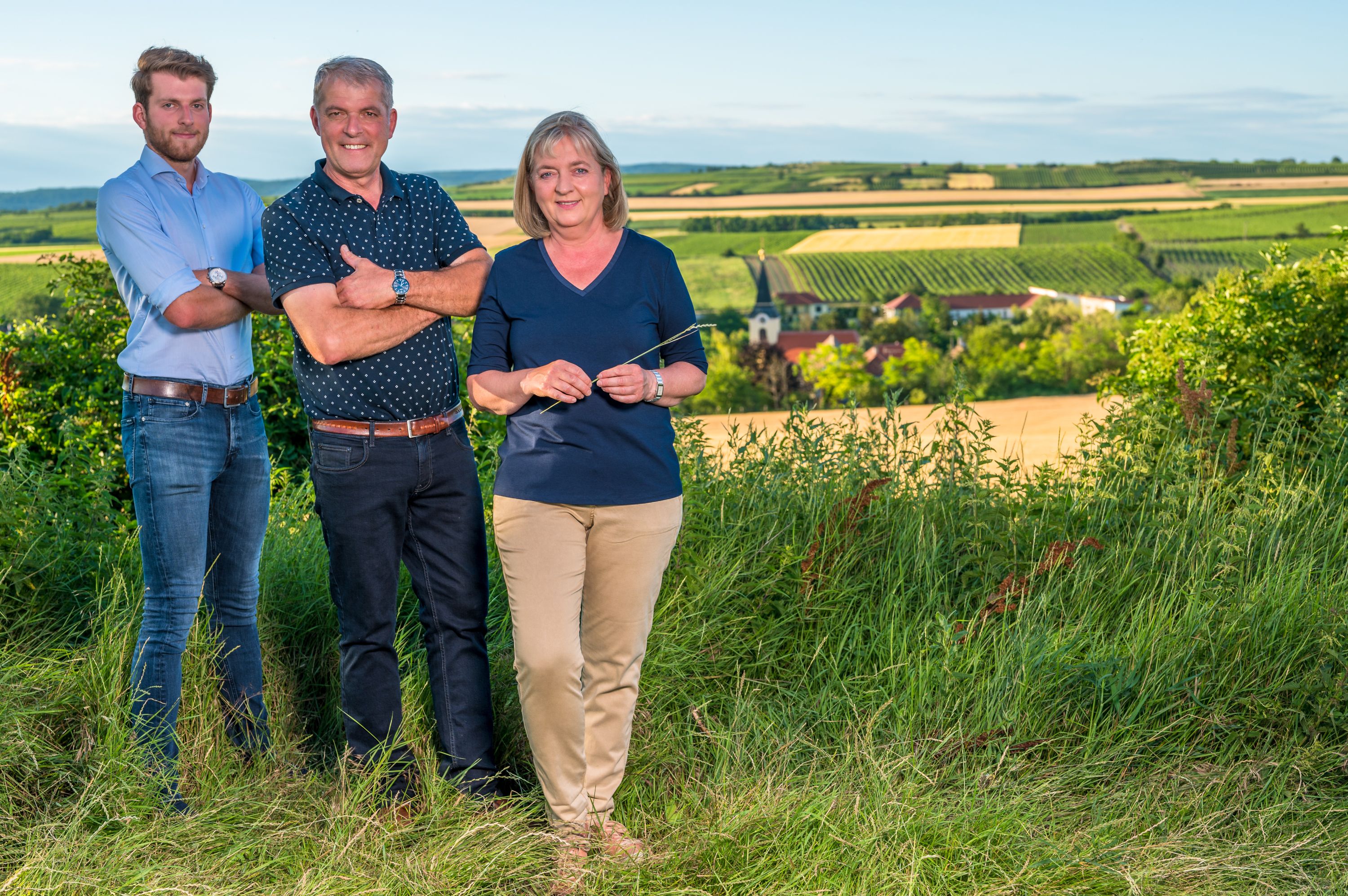 Tři lidé stojí na vinici s výhledem na venkovskou krajinu v pozadí.