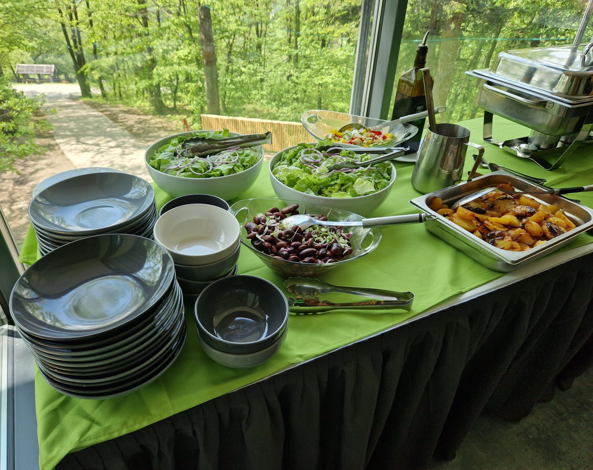 Venkovní bufet s různými saláty, talíři, bramborami a příbory na zeleném ubrusu.