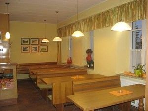 Pohled do interiéru útulné kavárny s dřevěnými stoly a lavicemi, vyzdobené obrázky a rostlinami.