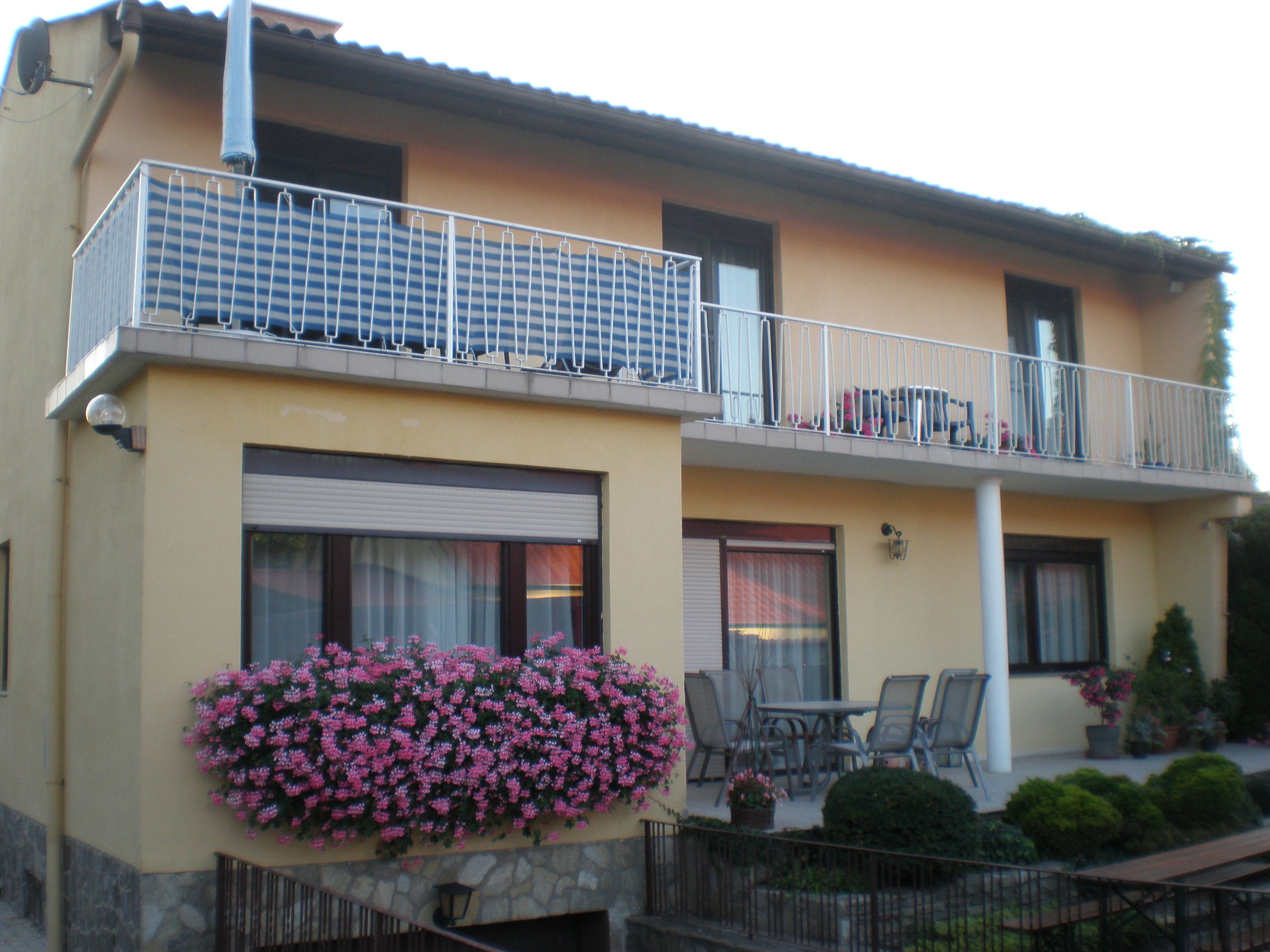 Dvoupodlažní dům s balkonem, květinami a zahradním nábytkem.