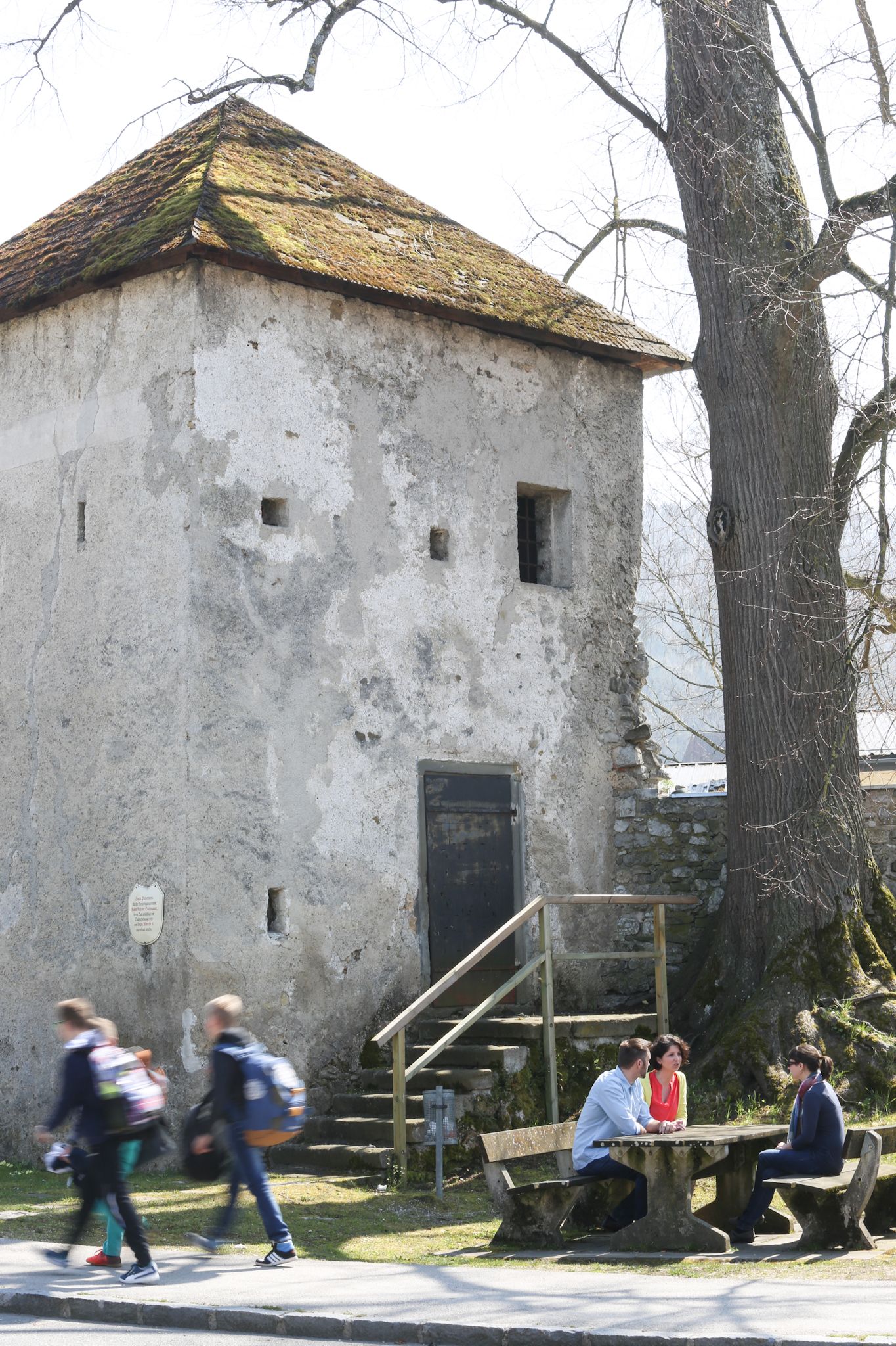 Stará věž se střechou porostlou mechem a stromem vedle ní. Tři lidé sedí u stolu, zatímco kolem procházejí dva lidé.