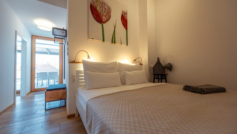 Moderní ložnice s manželskou postelí, světlou dřevěnou podlahou a uměleckými díly na stěnách. Dveře vedou na balkon s výhledem na střechy.