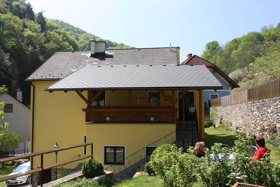 Žlutý dům s balkonem, obklopený zelení a kamennou zdí.