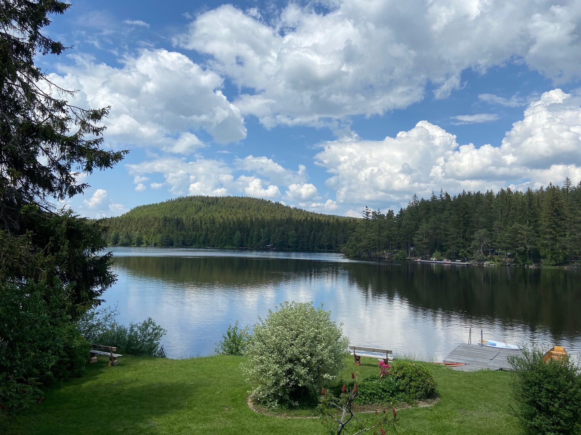 Klidné jezero se zalesněnými břehy a molem v popředí, obklopené zelenou krajinou a pod zataženou oblohou.