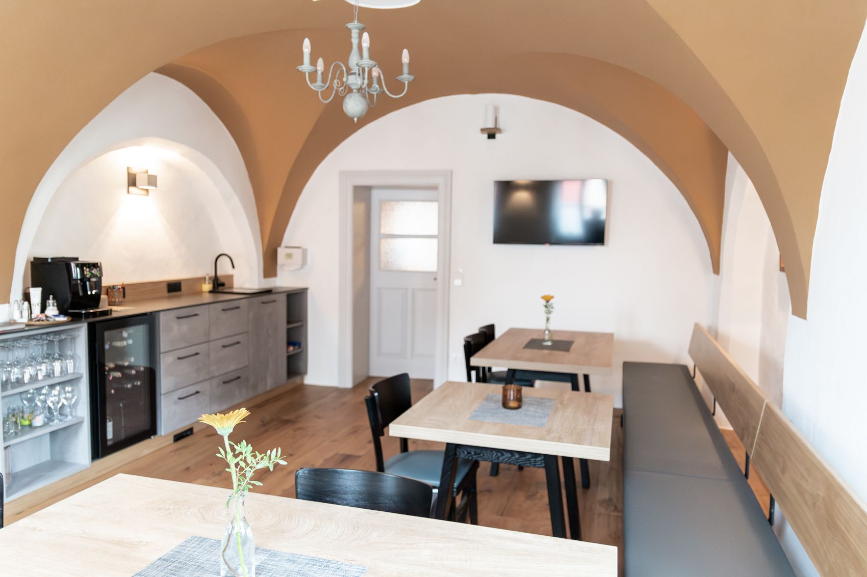 Útulná snídaňová místnost s dřevěnými stoly, židlemi a lavicí. Na stěně visí televize a k dispozici je kuchyňský kout s kávovarem.