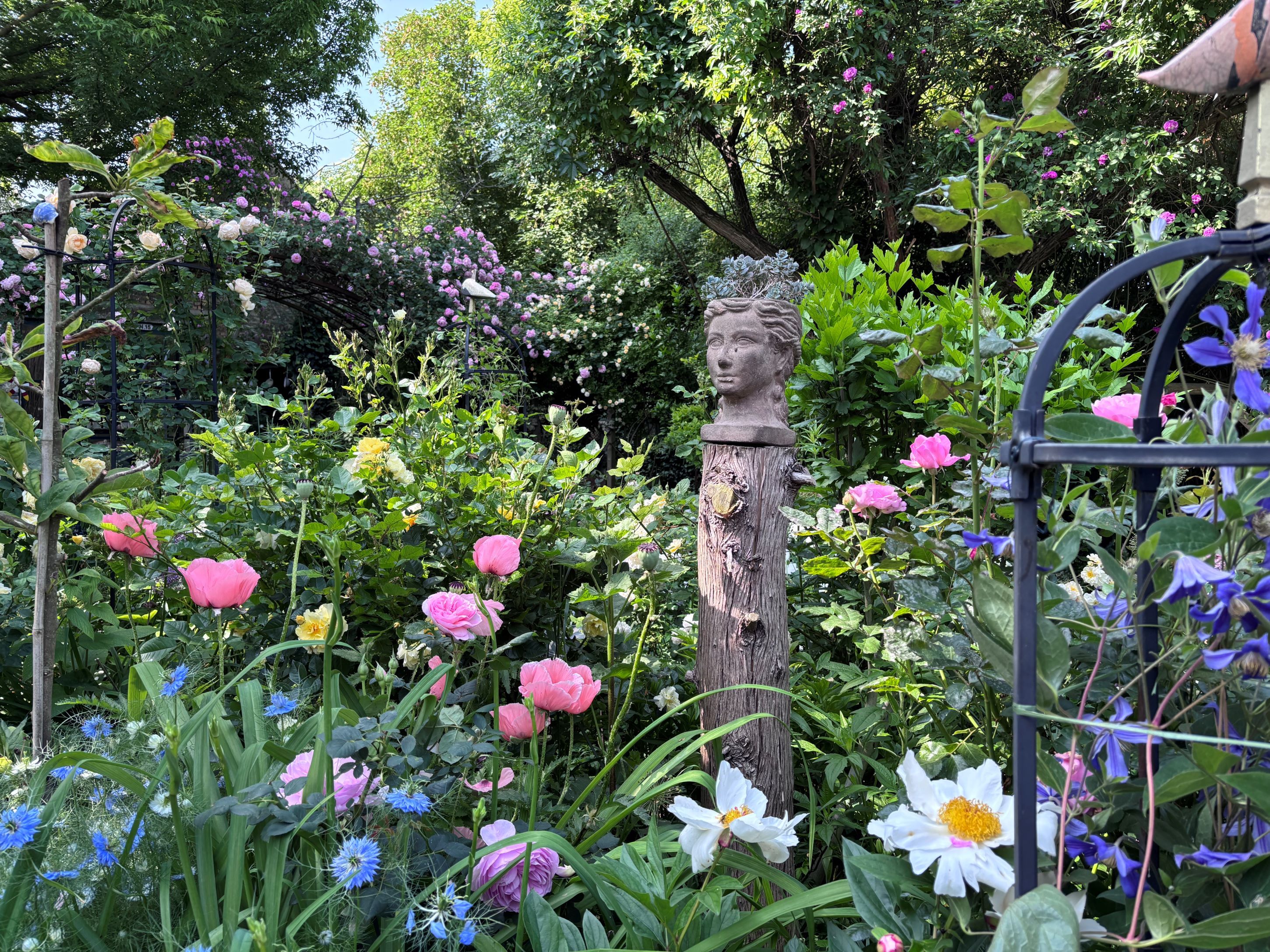Rozkvetlá zahrada s barevnými květy a bustou na dřevěném sloupku.