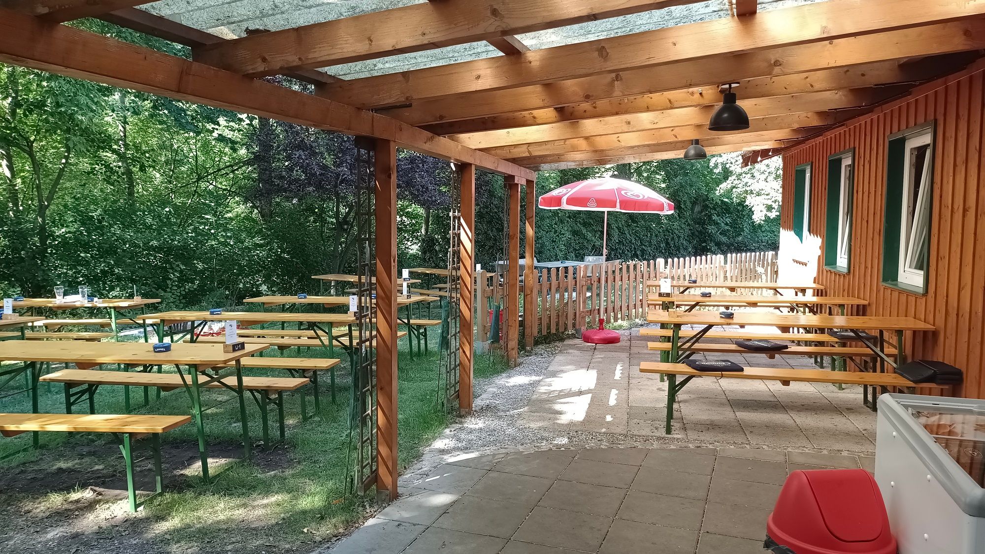 Krytá terasa s dřevěnými lavicemi a stoly, obklopená stromy a dřevěným plotem.