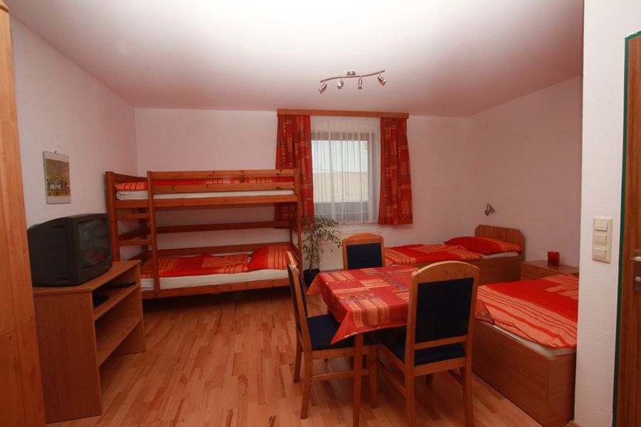 Jednoduše zařízený pokoj s palandou, samostatnou postelí, stolem a židlemi. Na polici je umístěna televize. V pozadí okno se závěsy.