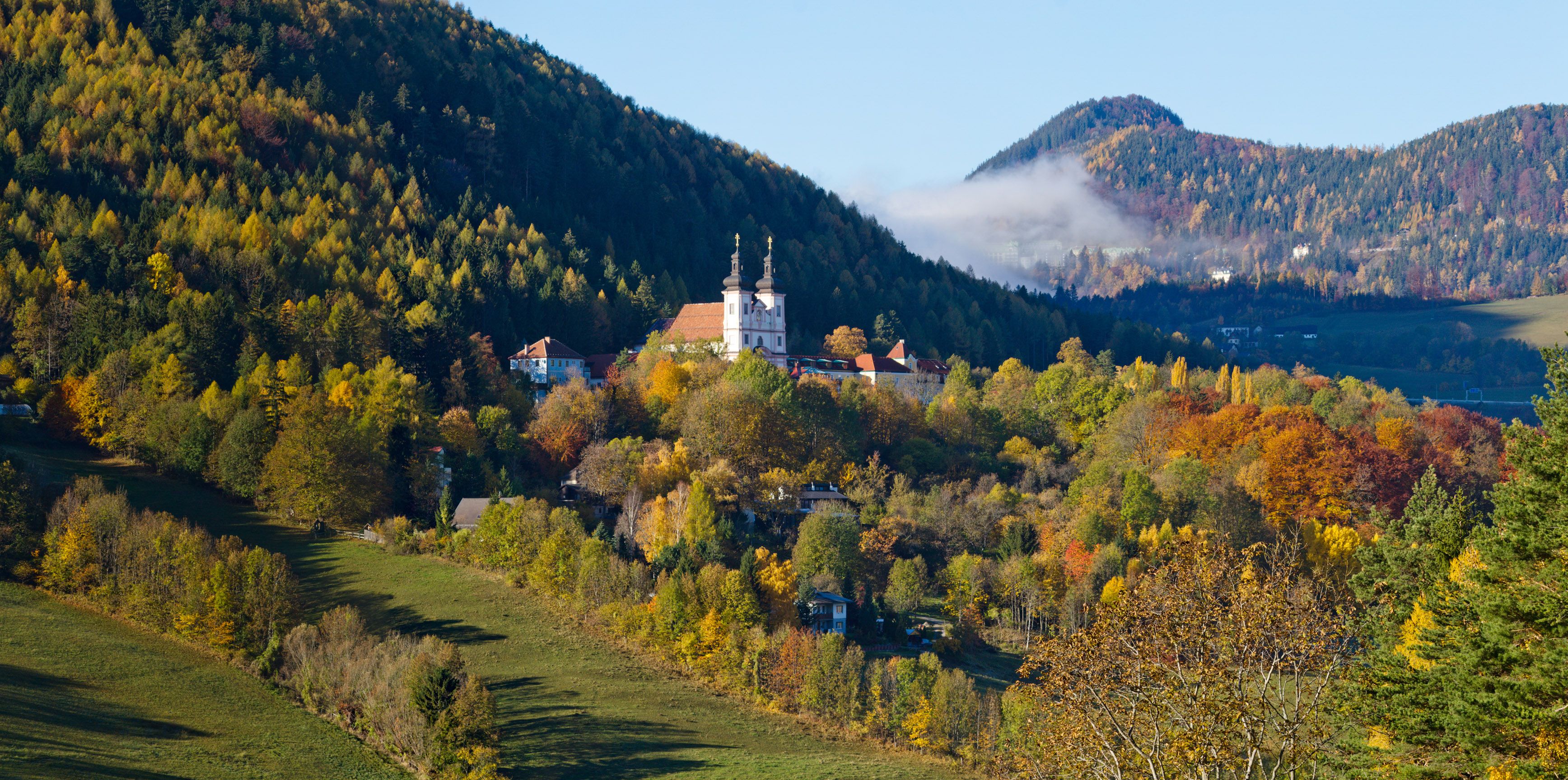 Podzimní krajina s kostelem v Maria Schutz, obklopená barevnými stromy a kopci.