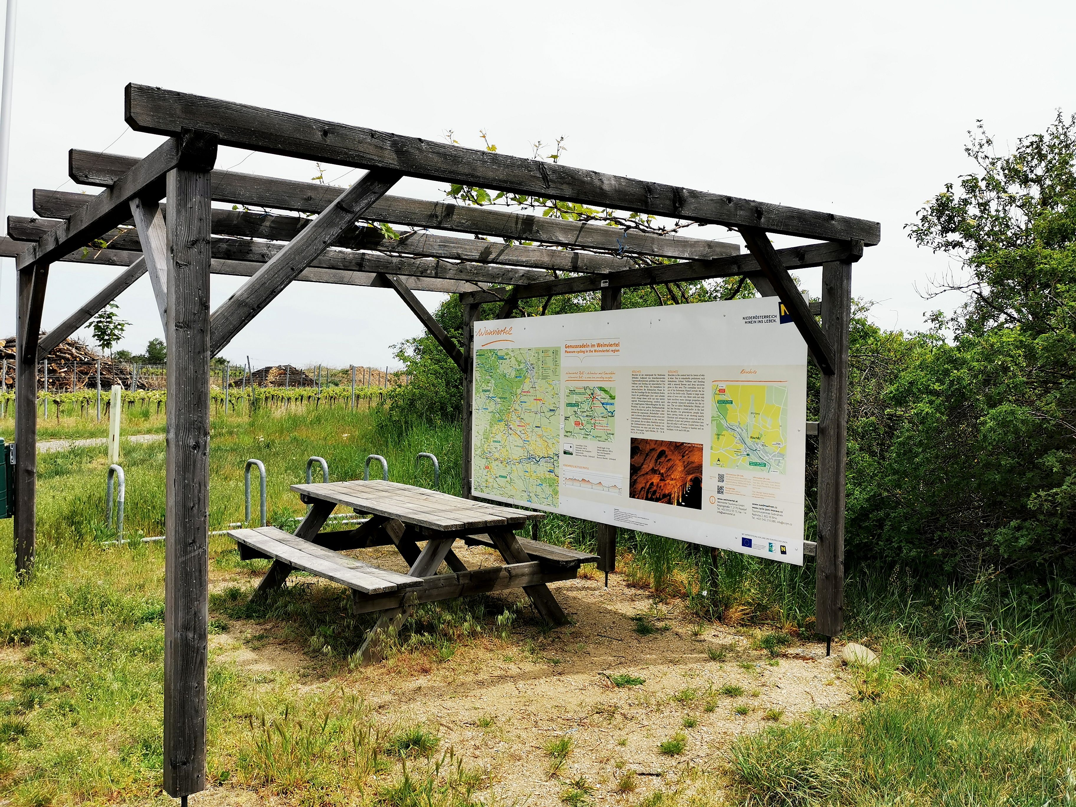 Dřevěná pergola s informační tabulí a piknikovým stolem ve venkovském prostředí.
