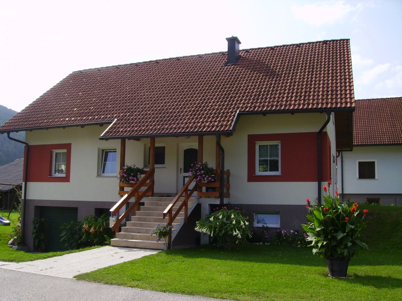 Samostatně stojící dům s červenou střechou a bílo-červenou fasádou, obklopený udržovanou zahradou a květinami.