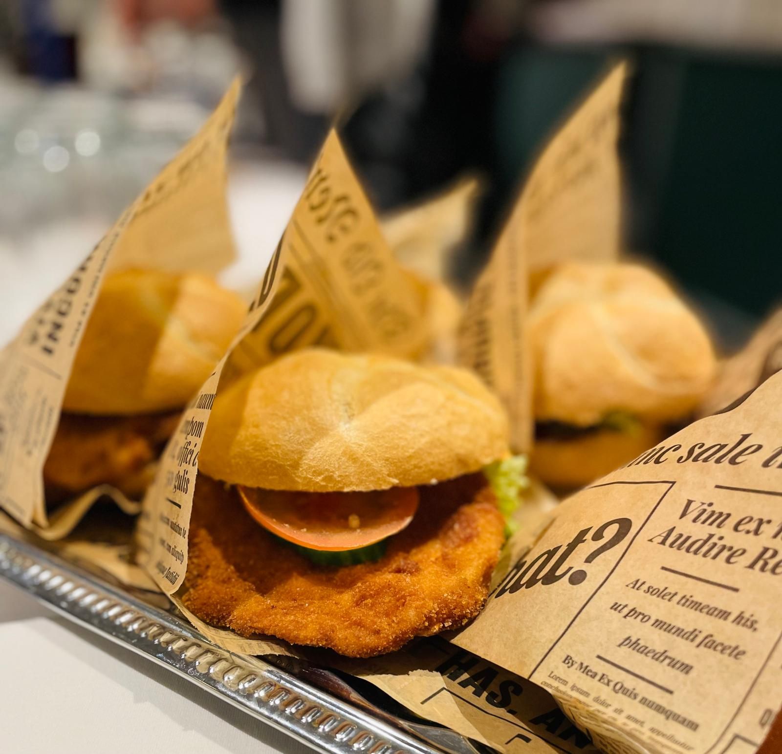 Několik hamburgerů zabalených v novinovém papíře na podnose, v popředí hamburger s obalovaným karbanátkem, rajčetem a okurkou.