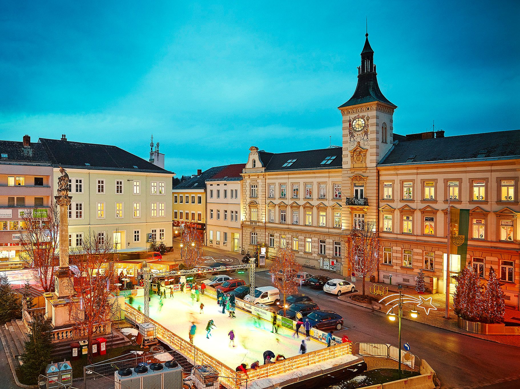 Kluziště ve městě za soumraku, obklopené historickými budovami a vánočním osvětlením.