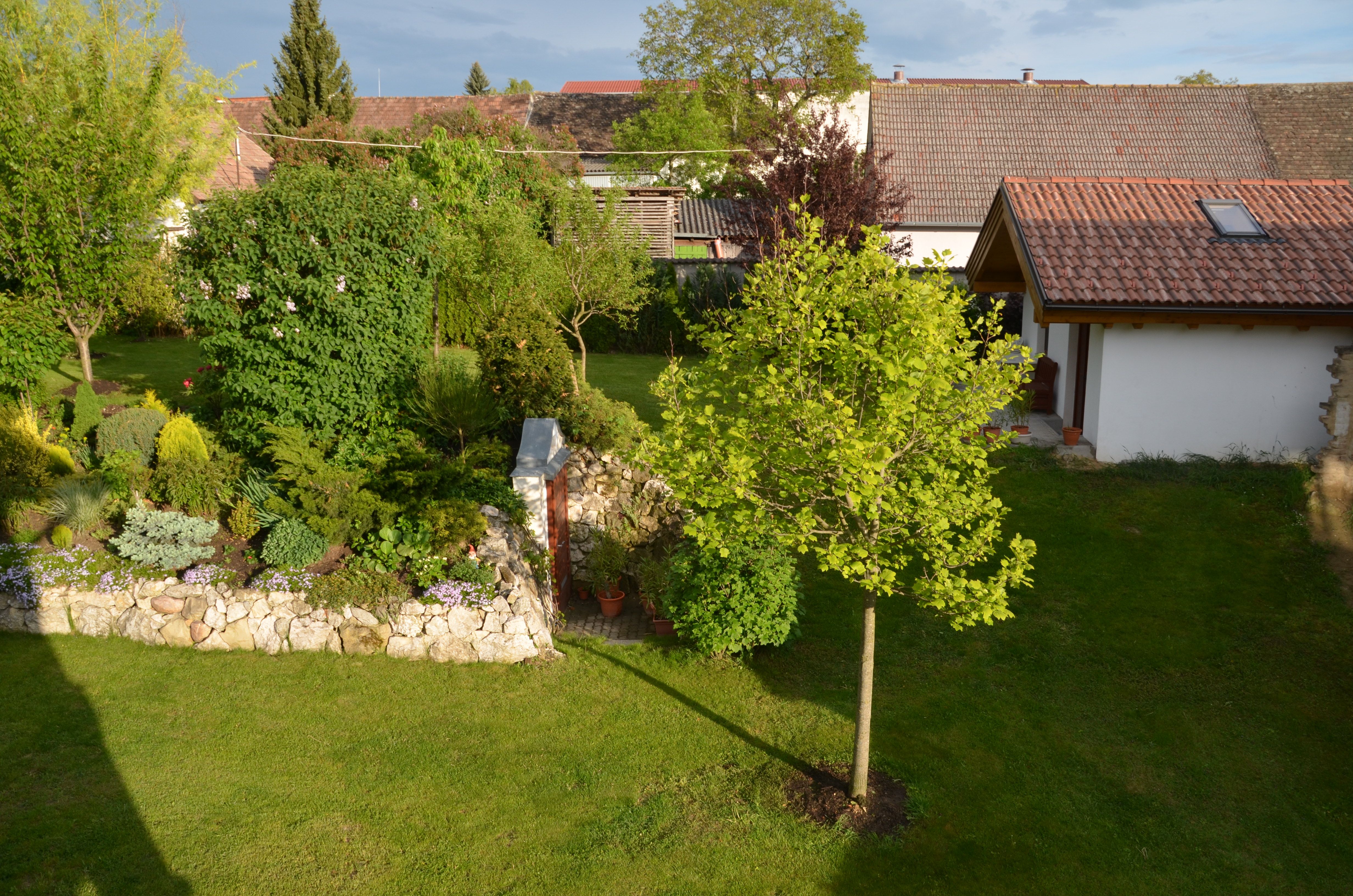 Udržovaná zahrada se stromy, keři a malou budovou s taškovou střechou.