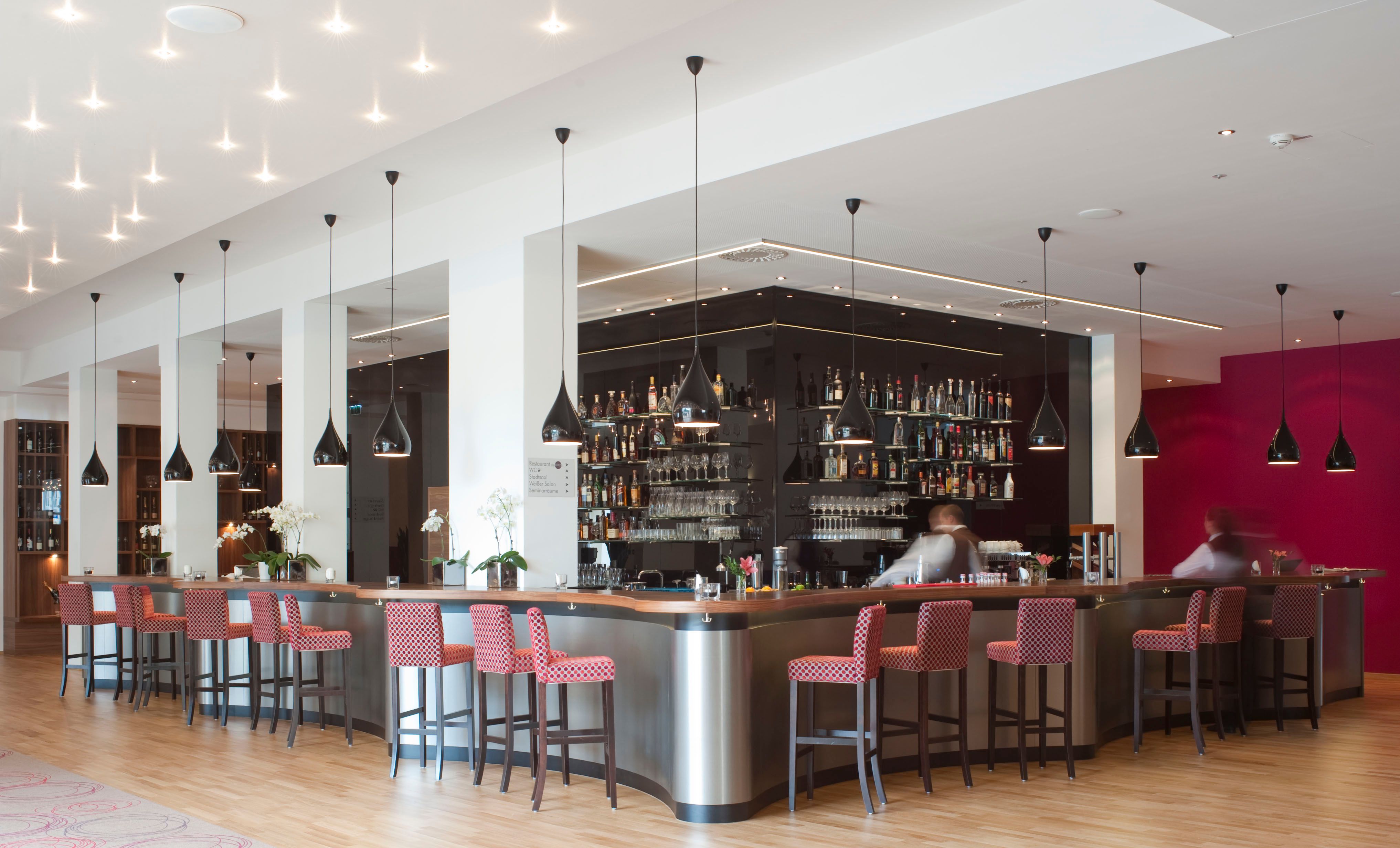 Moderní hotelový bar s červenými židlemi a černými závěsnými lampami.
