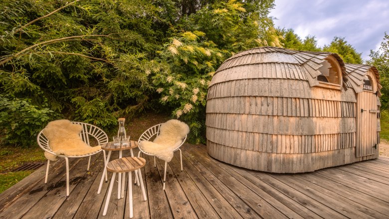 Dřevěná sauna se dvěma křesly a stolem na dřevěné terase obklopené zelení.