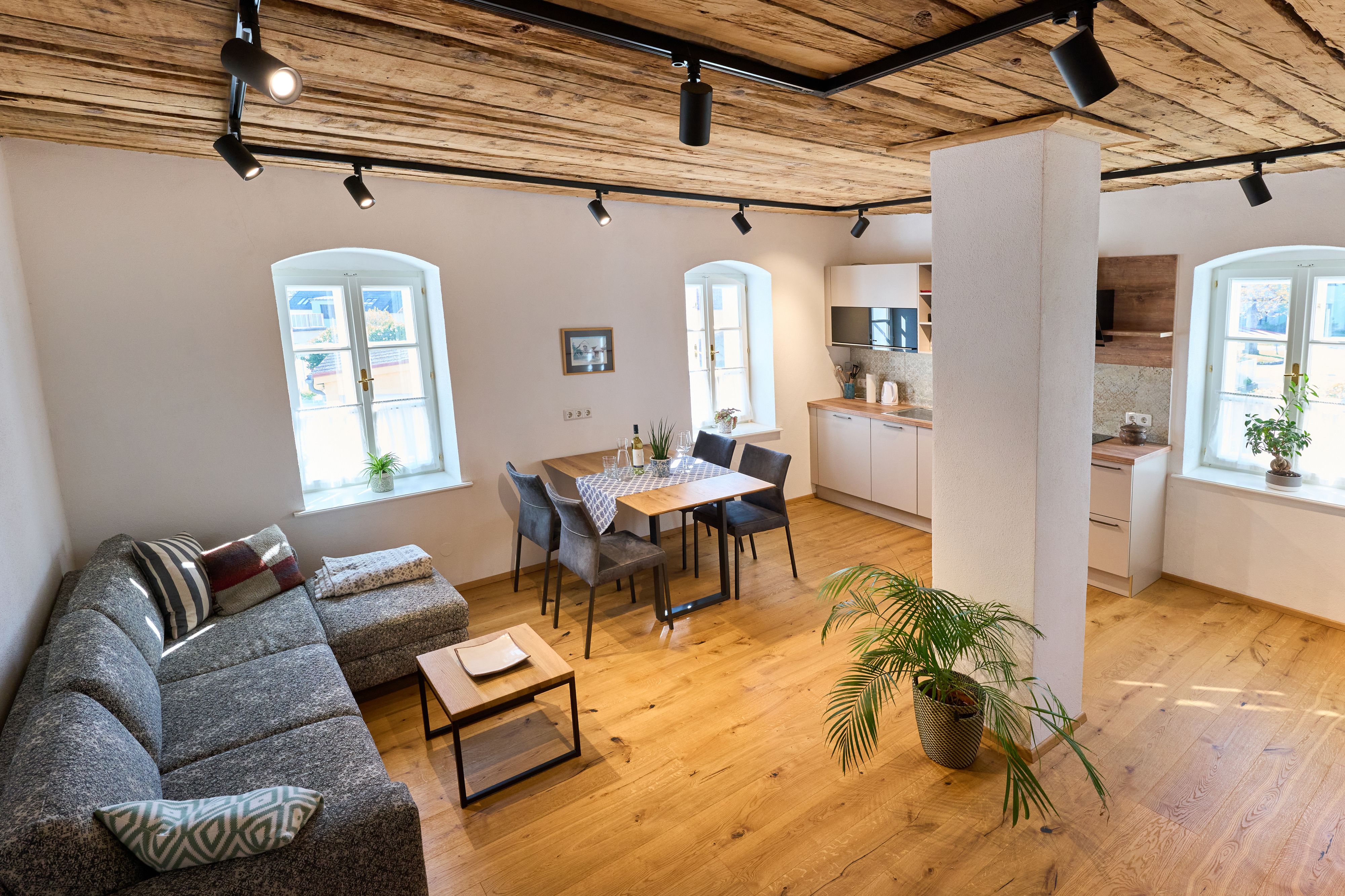 Moderní obývací pokoj s dřevěnou podlahou, šedou pohovkou, jídelním stolem a kuchyní.