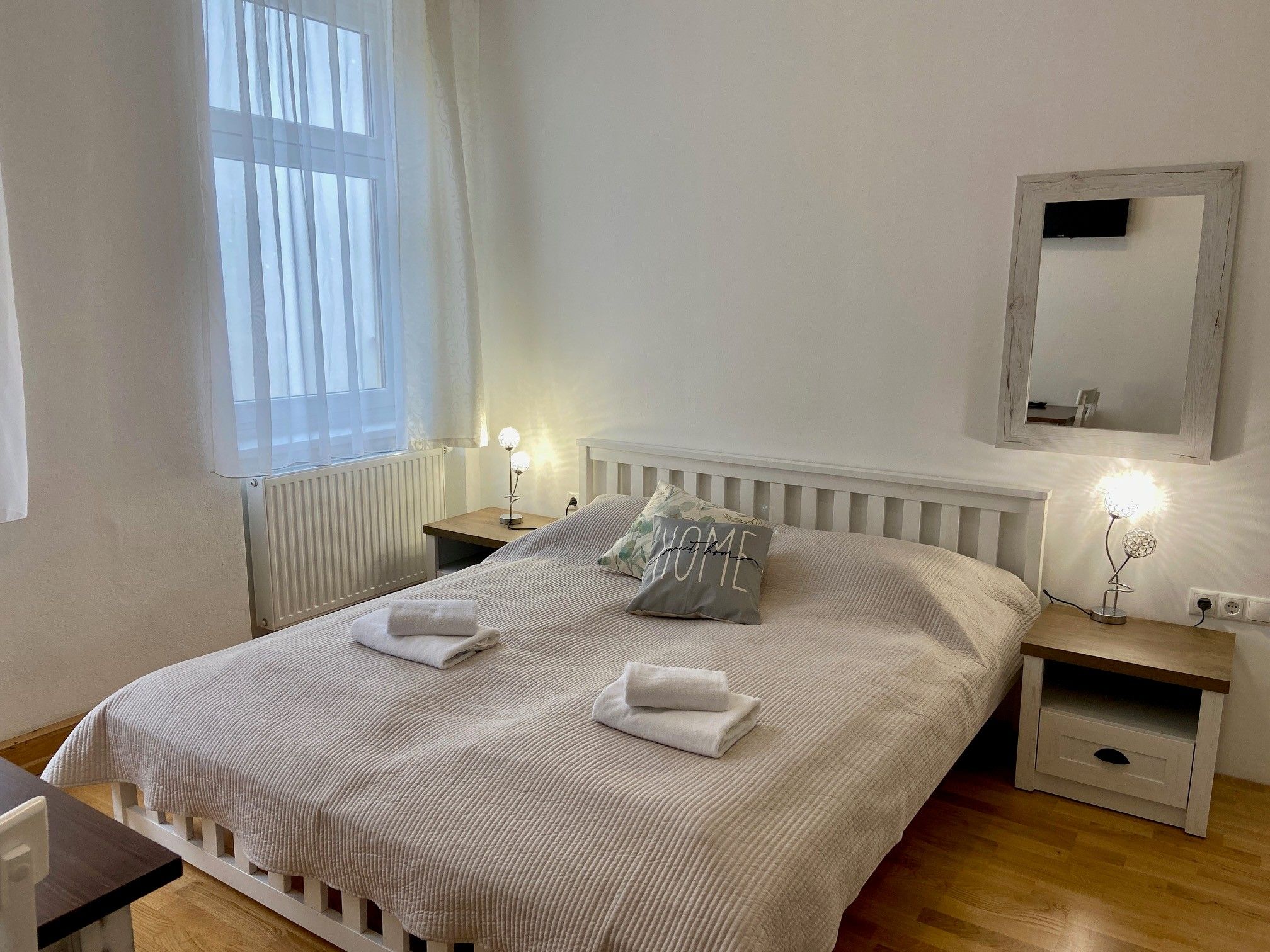 Útulný dvoulůžkový pokoj s velkou postelí, dvěma nočními stolky a zrcadlem na stěně. Pokoj je světlý a příjemný.