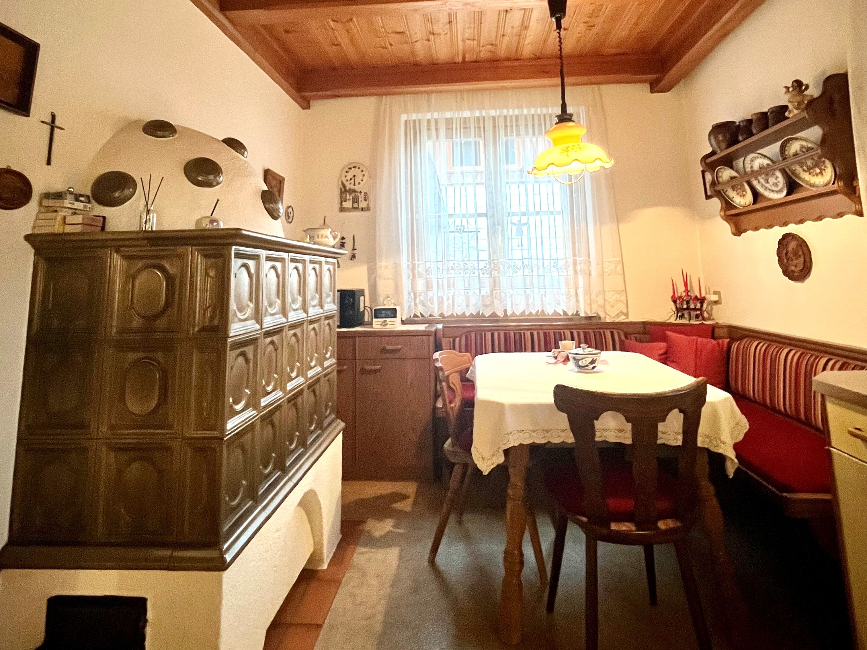 Útulná kuchyně s kachlovými kamny, dřevěným stolem a rohovou lavicí.