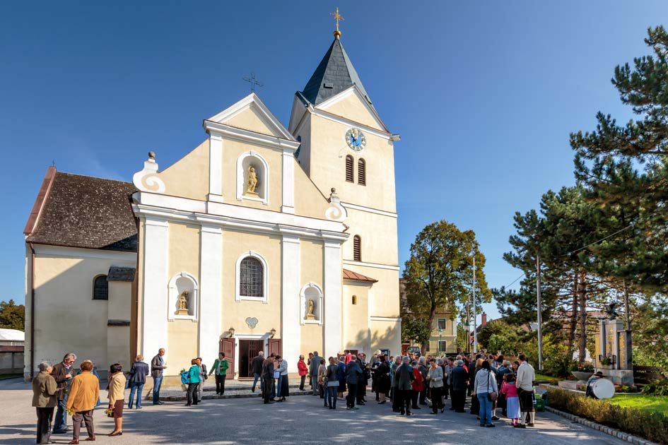 Lidé se za slunečného počasí shromažďují před kostelem s věží a hodinami.
