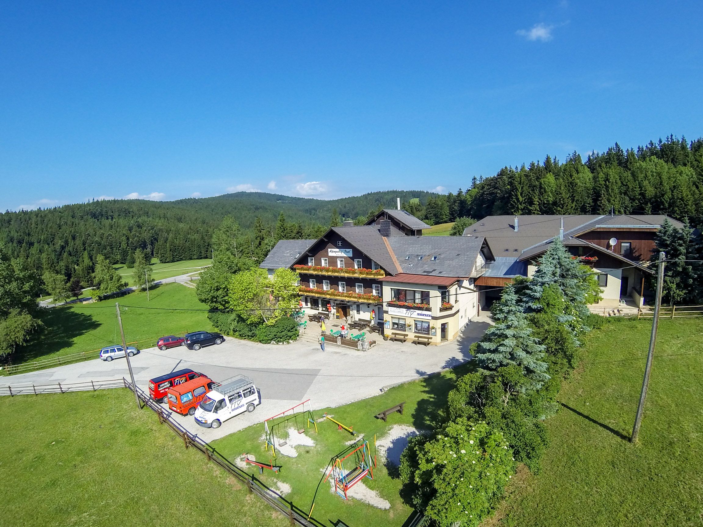 Alpengasthof Postl v zelené zalesněné krajině s několika zaparkovanými auty a dětským hřištěm v popředí.