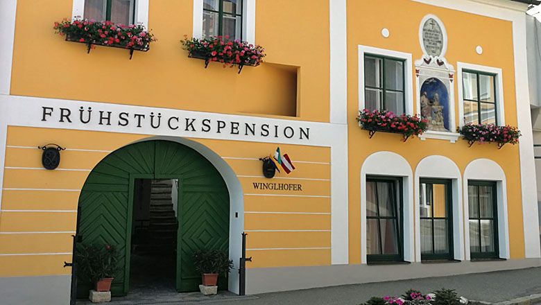 Žlutý dům pro hosty se zelenými dveřmi a květinovými truhlíky.