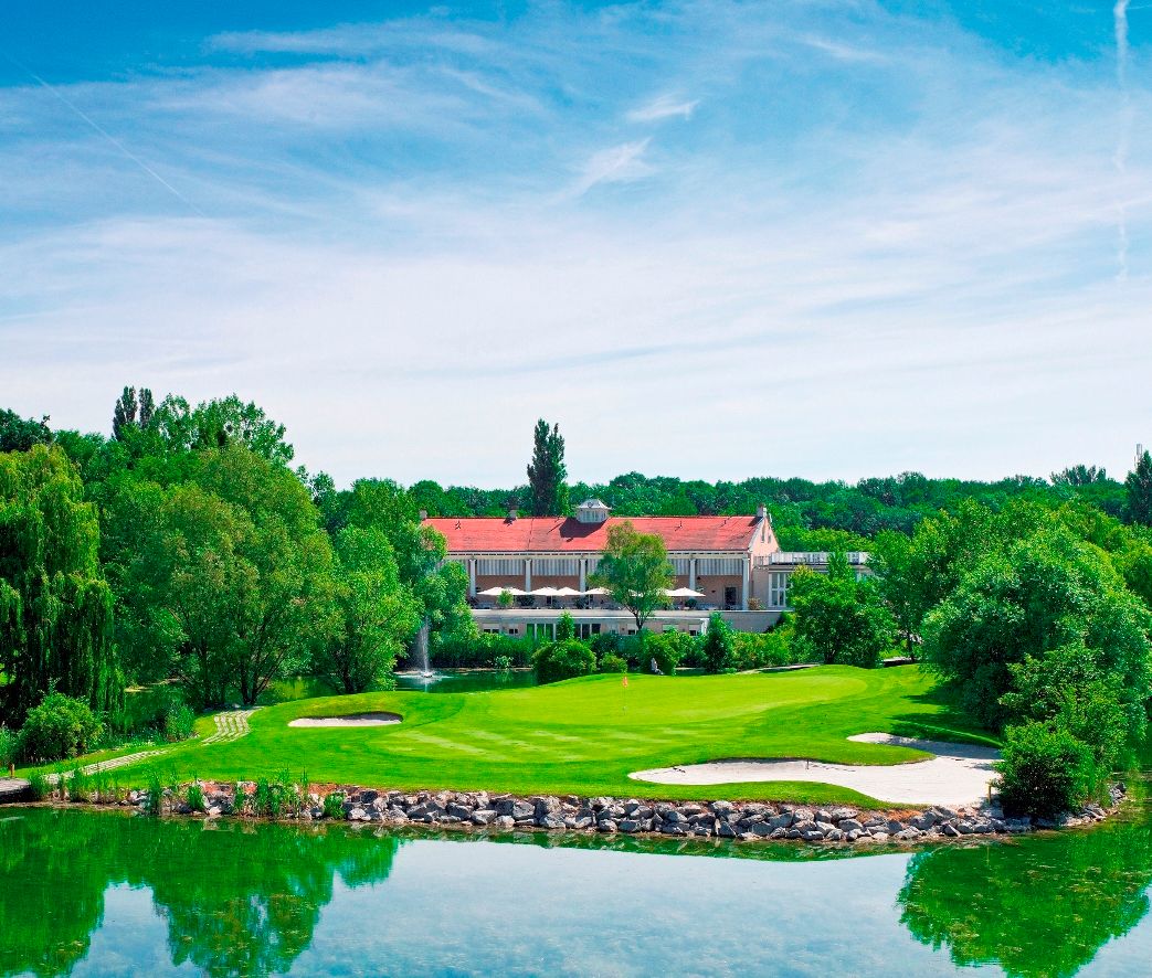 Golfové hřiště s klubovnou v pozadí, obklopené stromy a rybníkem.